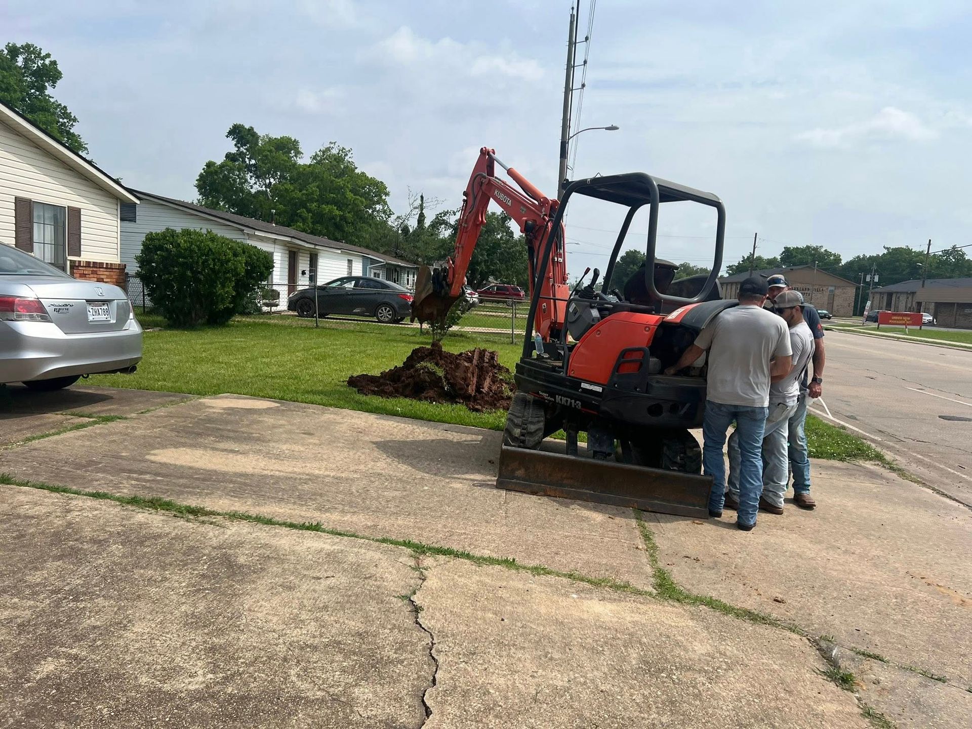 Two workers stand by an orange excavator loading dirt beside a suburban street and houses.