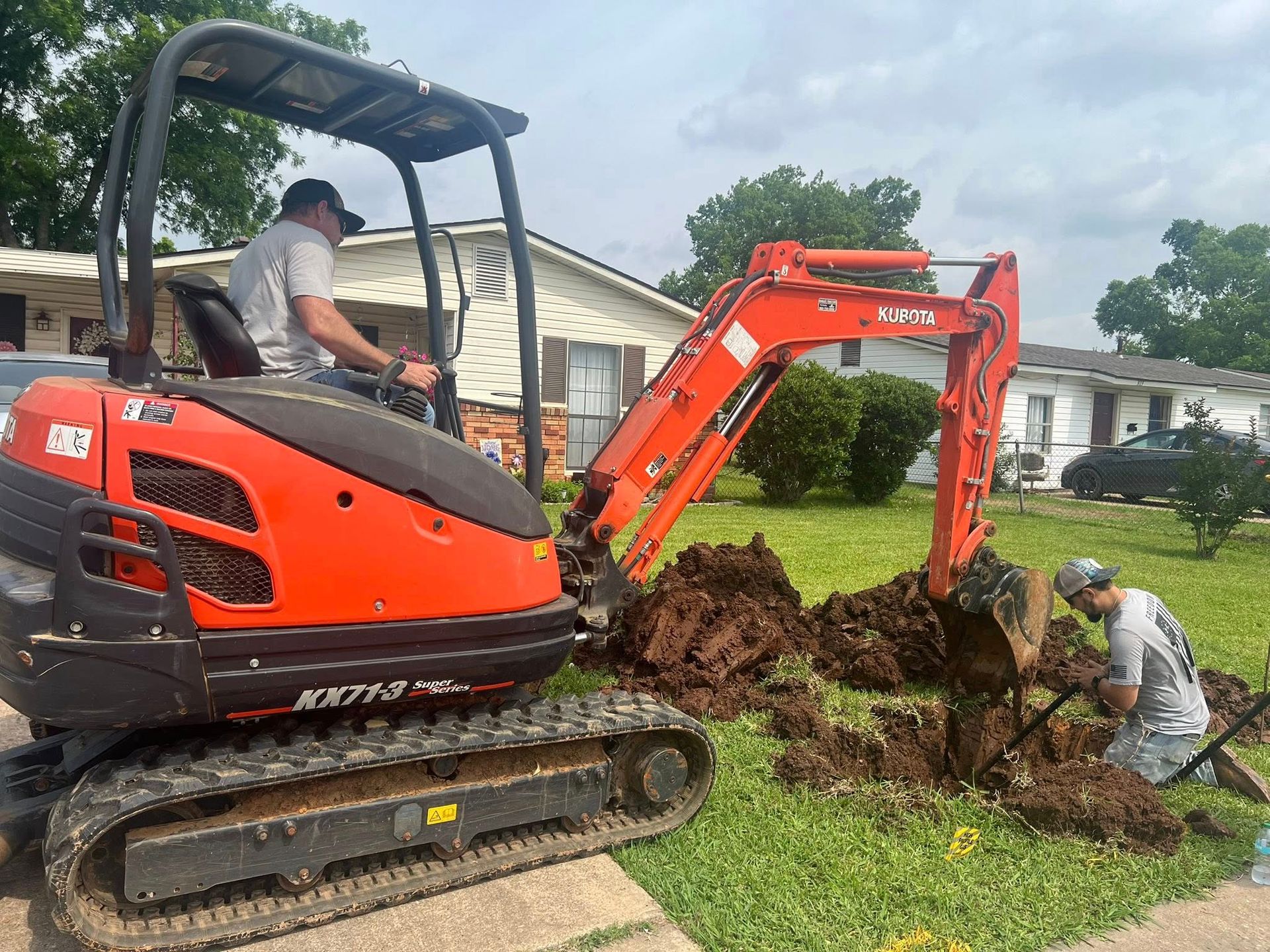 Orange excavator digging a trench in a grassy yard while workers clear soil