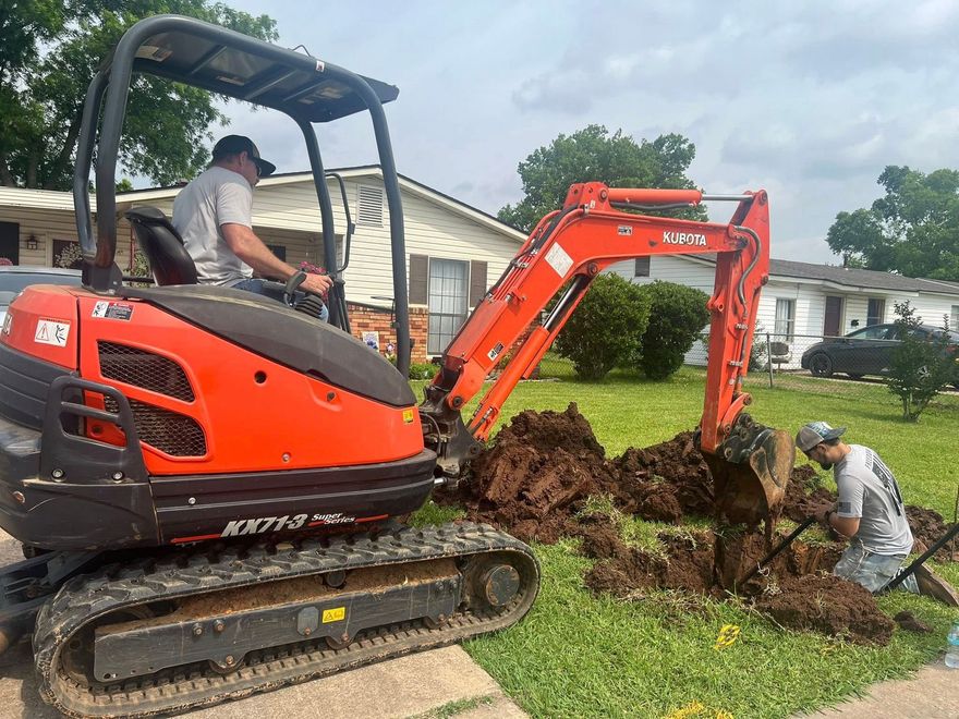 Orange excavator digging a trench in a grassy yard while workers clear soil