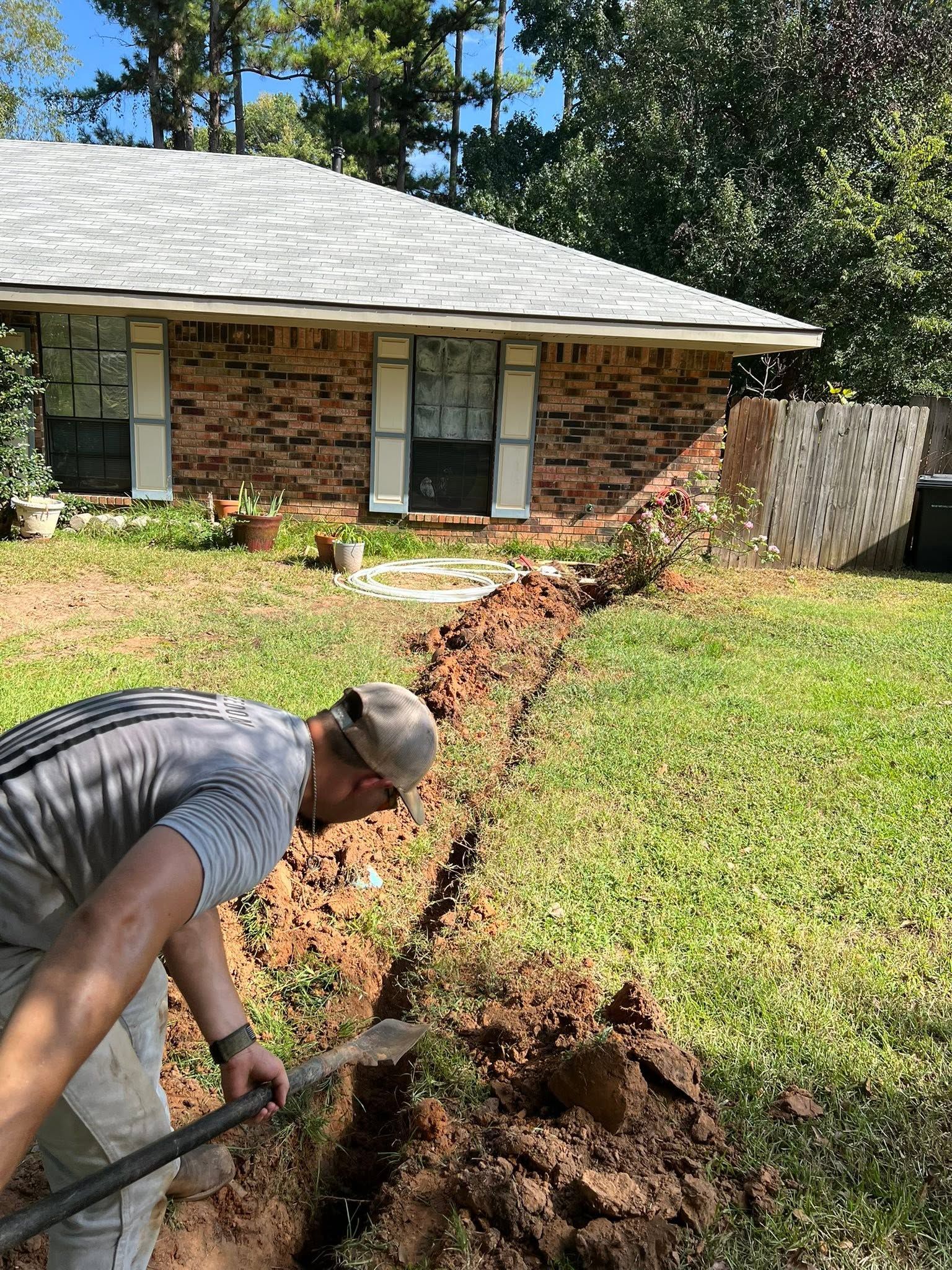 Person digging a trench in a front yard beside a brick house on a sunny day