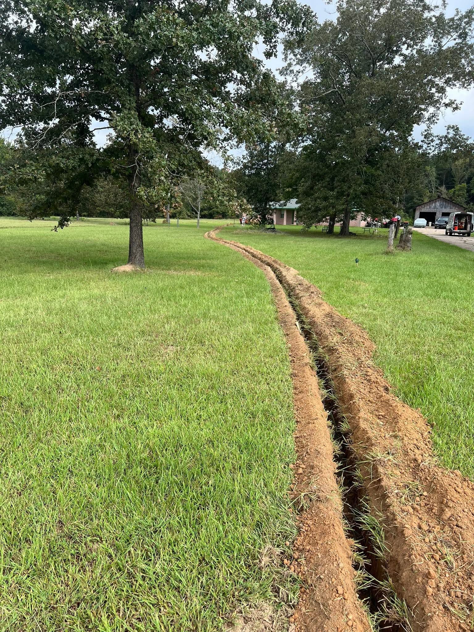 Grassy yard with a shallow dirt trench running toward trees and parked cars in the distance