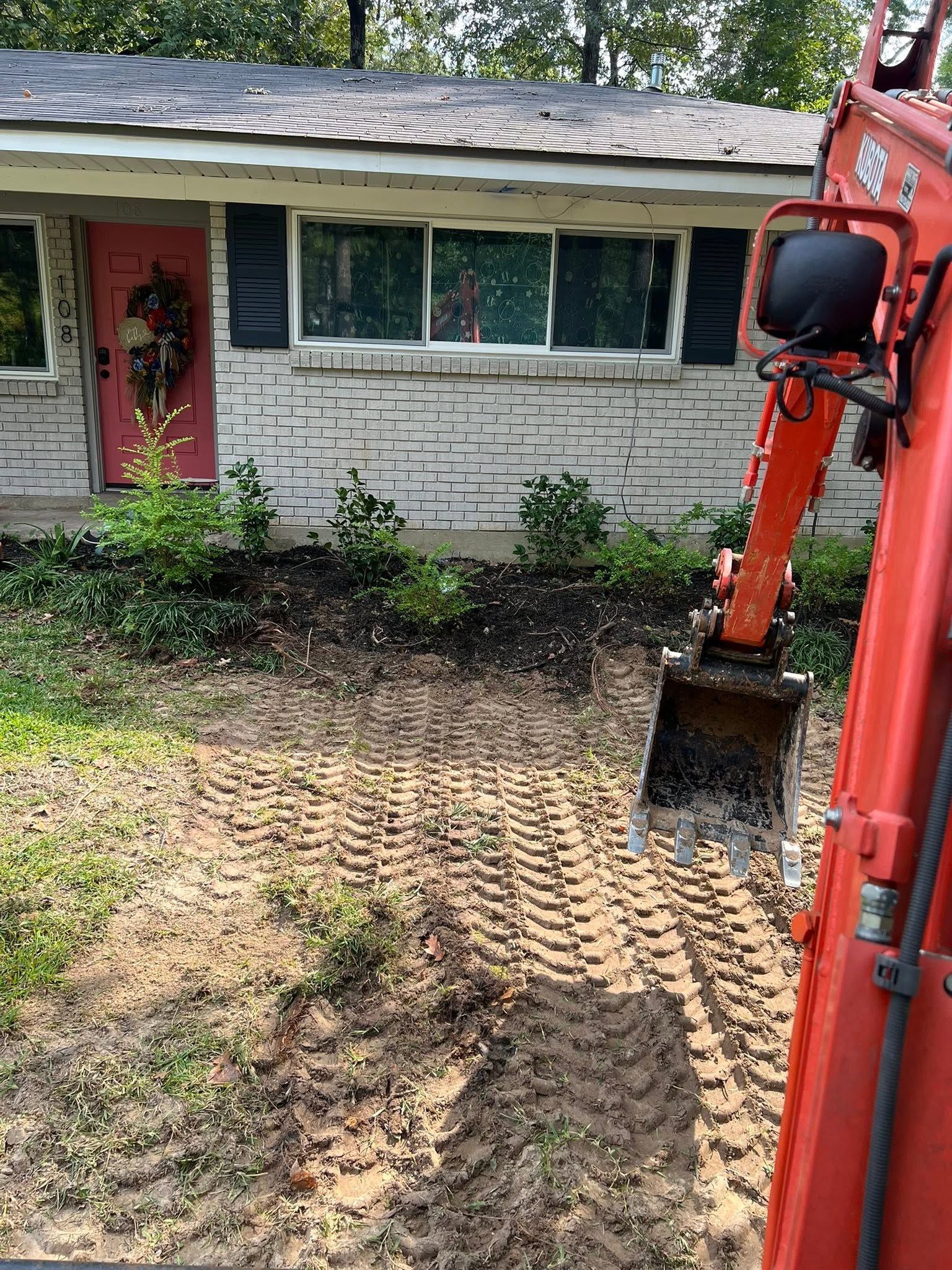 Backyard with a red excavator beside a house, digging up muddy ground near the foundation