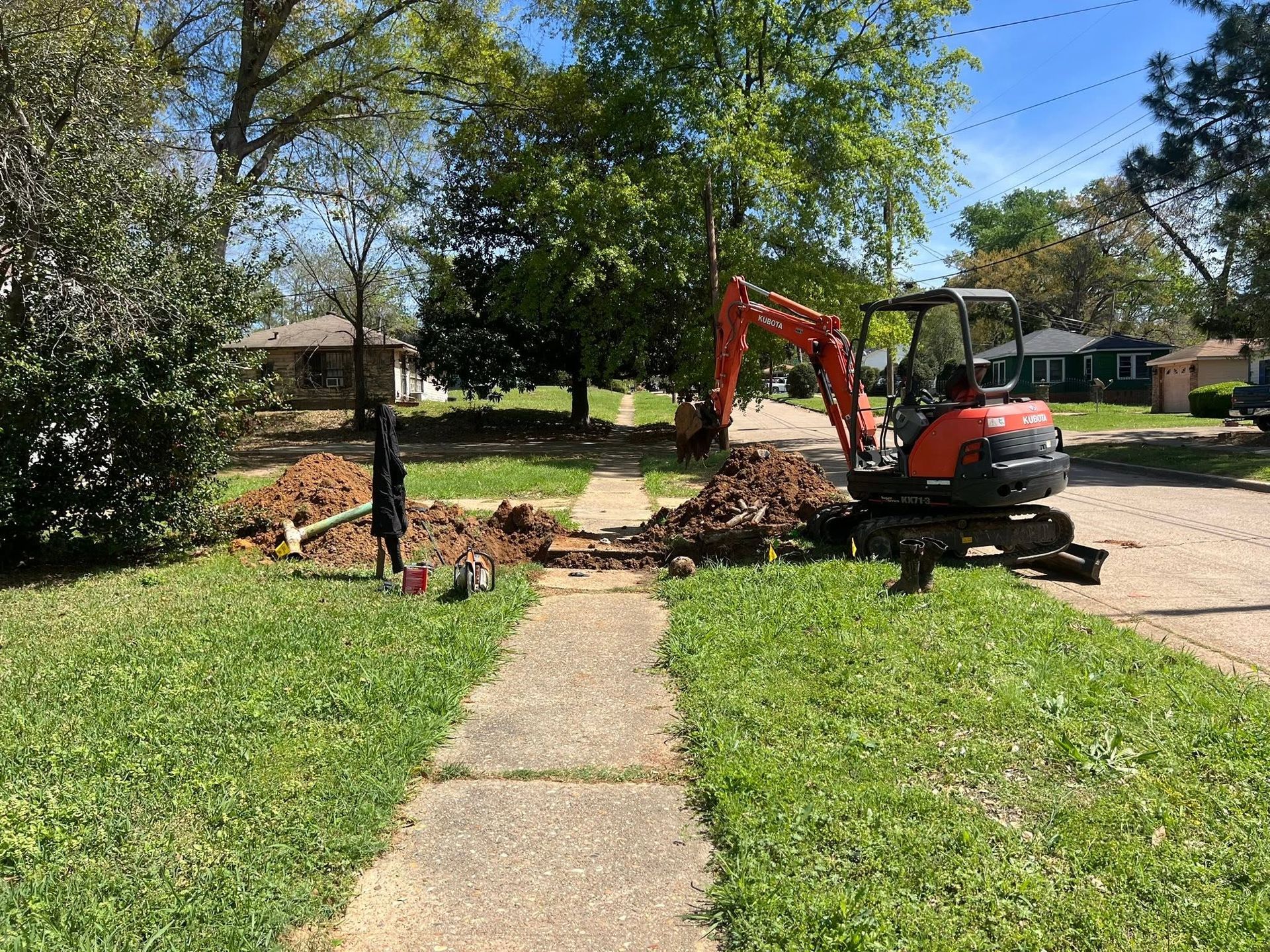 Orange mini excavator clearing a fallen tree beside a sidewalk in a sunny residential yard