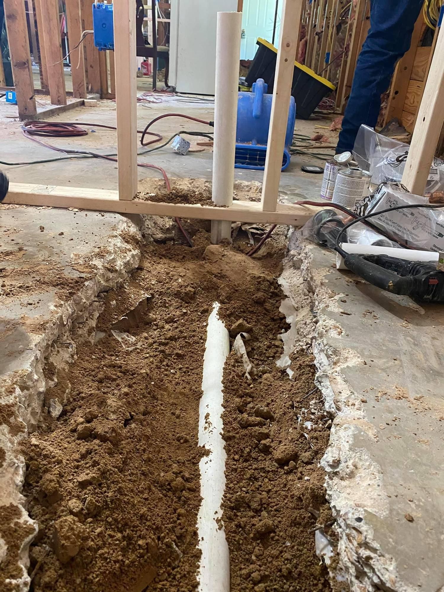 Construction trench with exposed white pipe running through dirt beside a damaged concrete slab and wall framing.