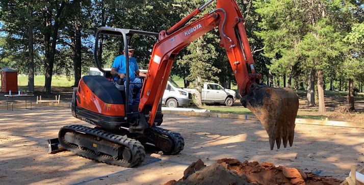 Orange excavator digging soil in a park, with a worker in blue inside the cab.