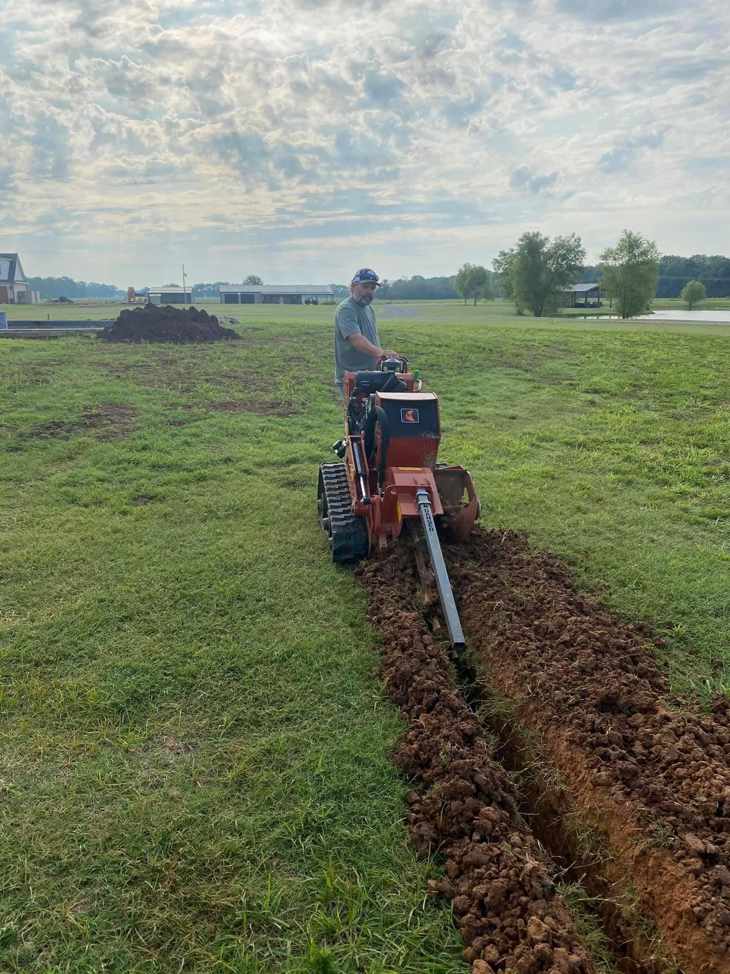Person operating a small tractor cutting a trench through a grassy field under a cloudy sky