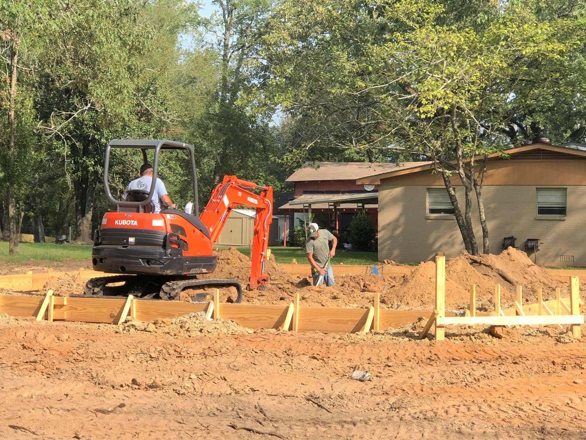 Orange excavator at a dirt construction site beside a house, with two workers nearby.