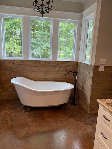 Freestanding white bathtub beneath windows in a warm-toned bathroom with a chandelier and wood-look tile floor