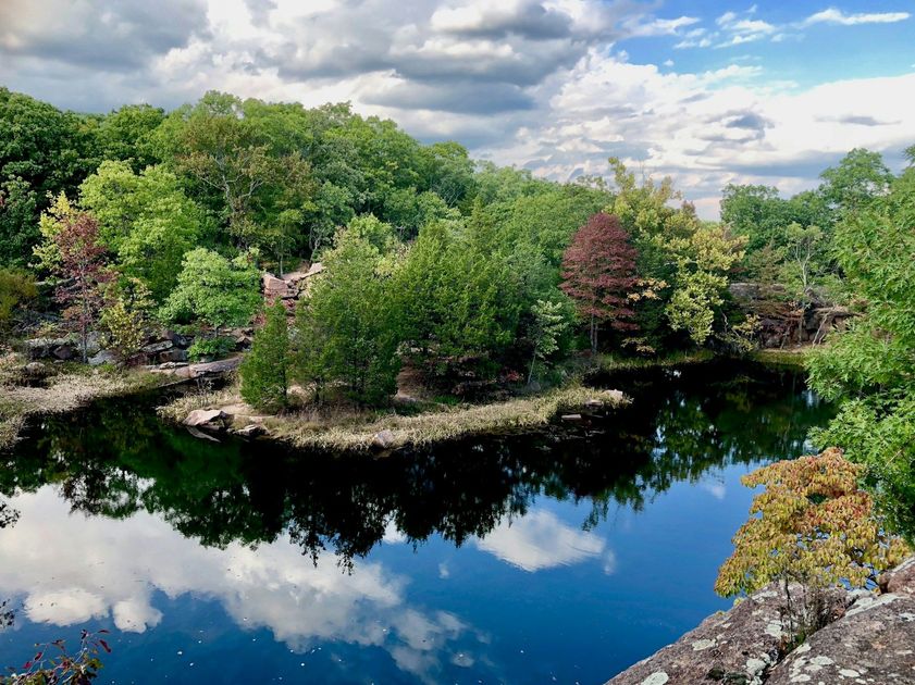 Serene pond reflecting clouds, surrounded by lush green trees and rocky shoreline under a blue sky