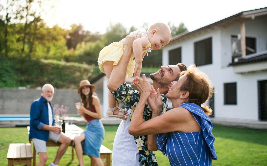 Outdoor family gathering with a baby being lifted by an adult in a backyard near a white house