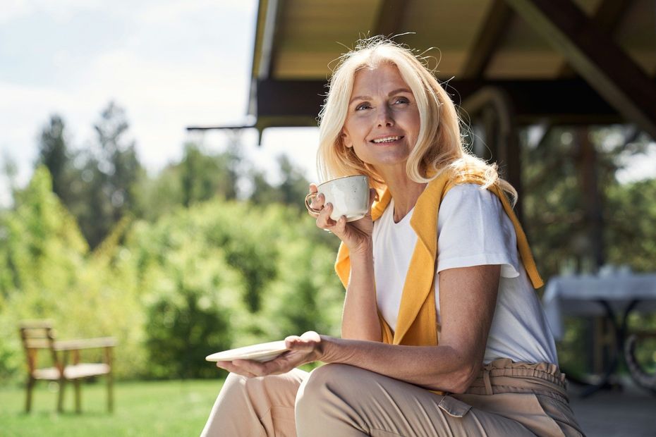 Woman sitting outdoors, smiling and holding a cup and plate on a sunny patio.