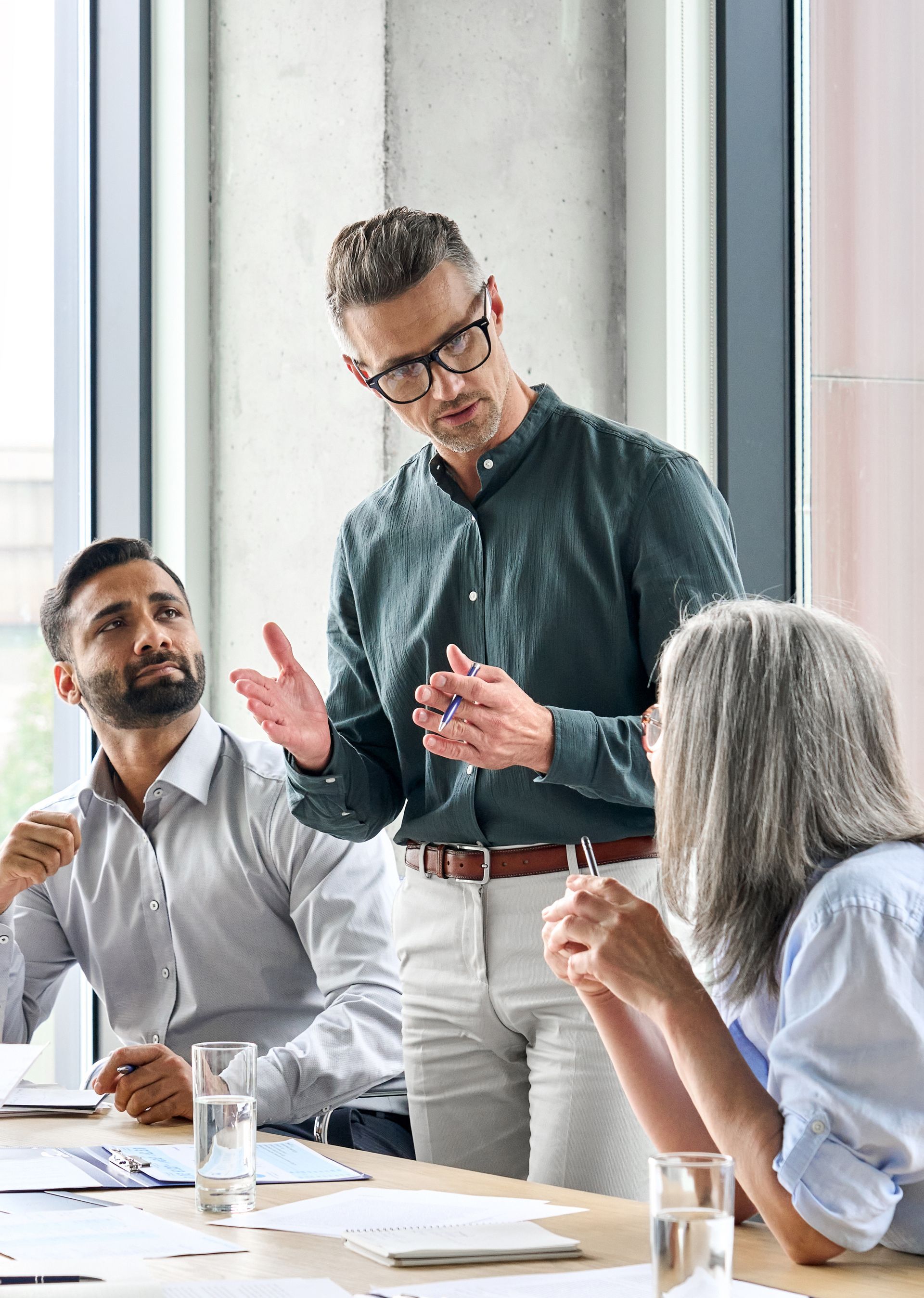 Three coworkers discussing in a bright meeting room, one standing and speaking while two listen at a table.