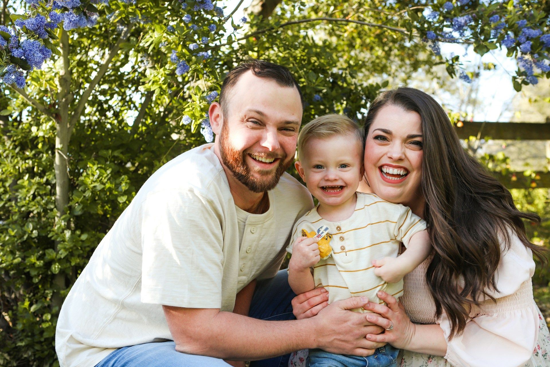 Smiling family of three sitting outdoors in a sunny garden, with trees and flowers behind them