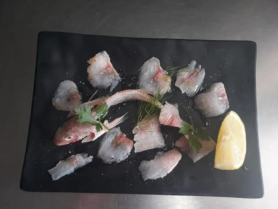 Fettine di pesce crudo su un piatto nero con erbe aromatiche e uno spicchio di limone.