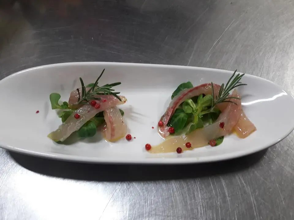 Due antipasti di pesce serviti su un piatto bianco con verdure, erbe aromatiche e salsa rossa.