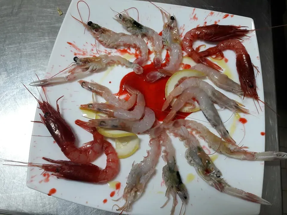 Gamberi e gamberetti crudi assortiti disposti su un tagliere bianco con salsa rossa e fette di limone.