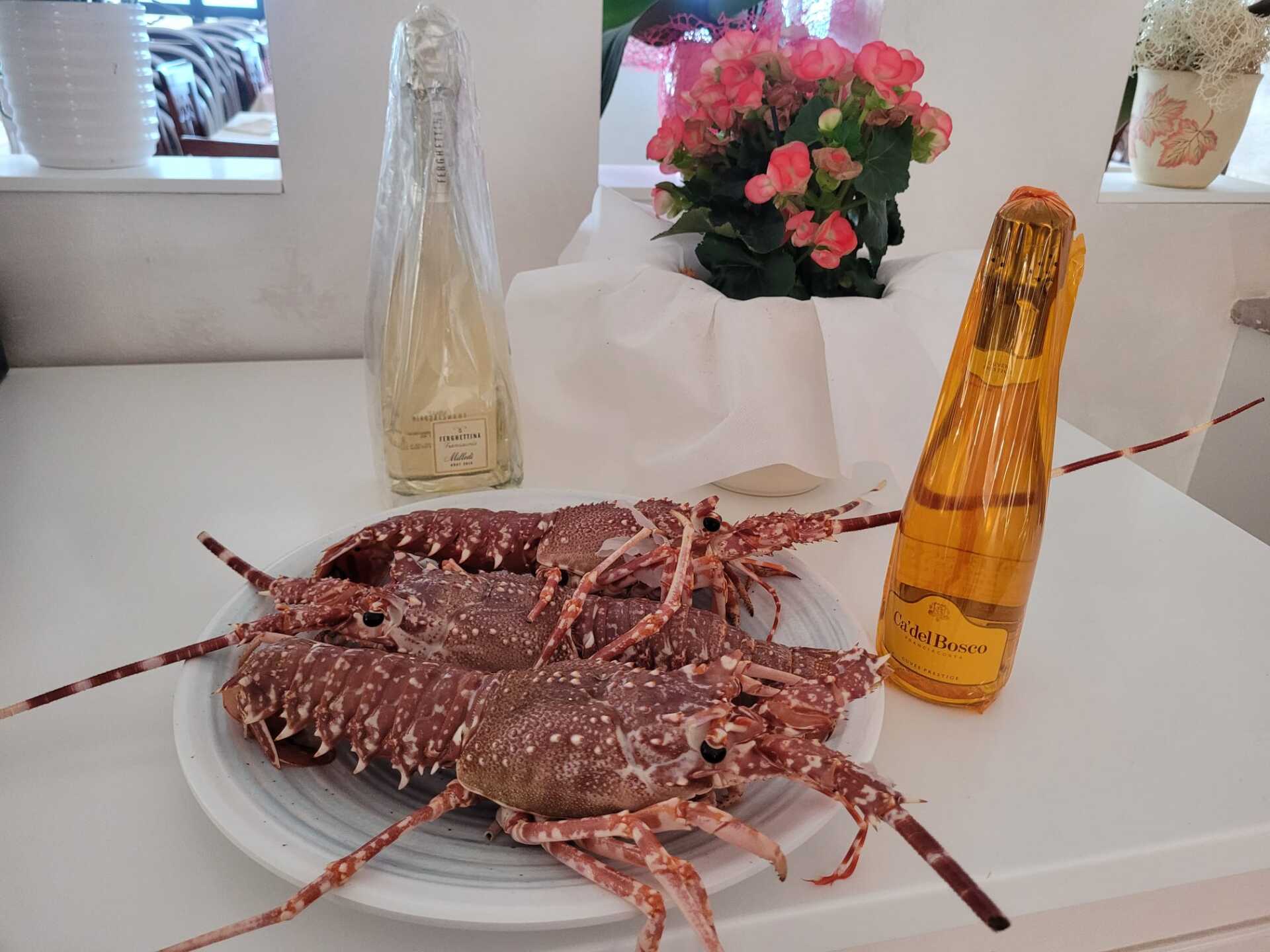 Un piatto di aragoste cotte su un tavolo con bottiglie e una composizione floreale nelle vicinanze.