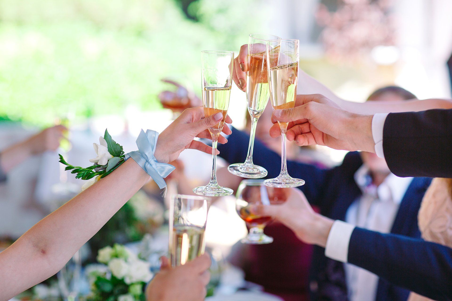 Mani che brindano con calici di champagne durante una festa all'aperto, con gli ospiti sfocati sullo sfondo.