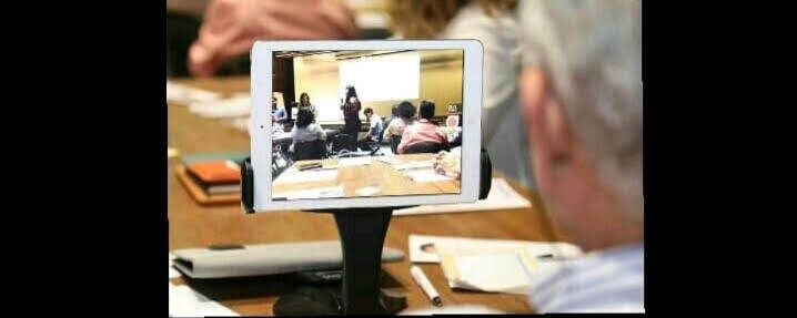 Um tablet sobre um suporte exibe uma reunião. Uma pessoa de cabelos grisalhos está em primeiro plano.