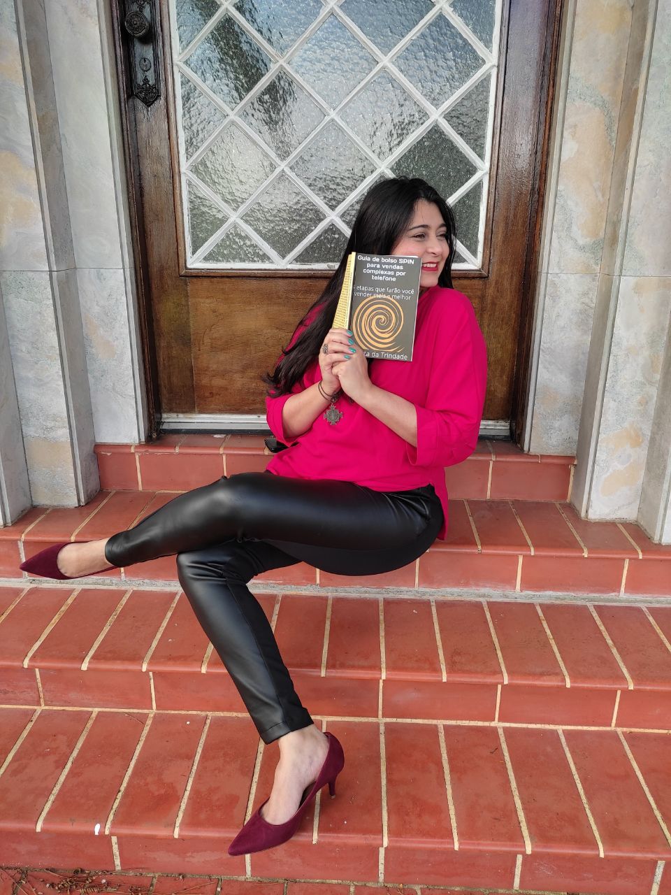 Mulher sentada em degraus de tijolo, segurando um livro, vestindo uma blusa rosa, calça preta e sapatos de salto alto cor vinho.