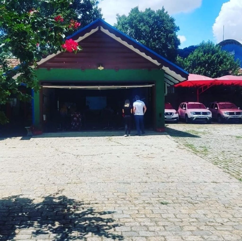 Garagem verde com a porta aberta, duas pessoas do lado de fora. Carros rosa estacionados nas proximidades.