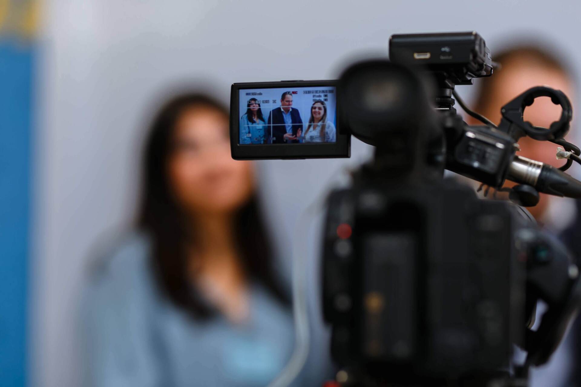 Câmera de vídeo gravando três pessoas durante uma entrevista. Mulher em primeiro plano, fora de foco.