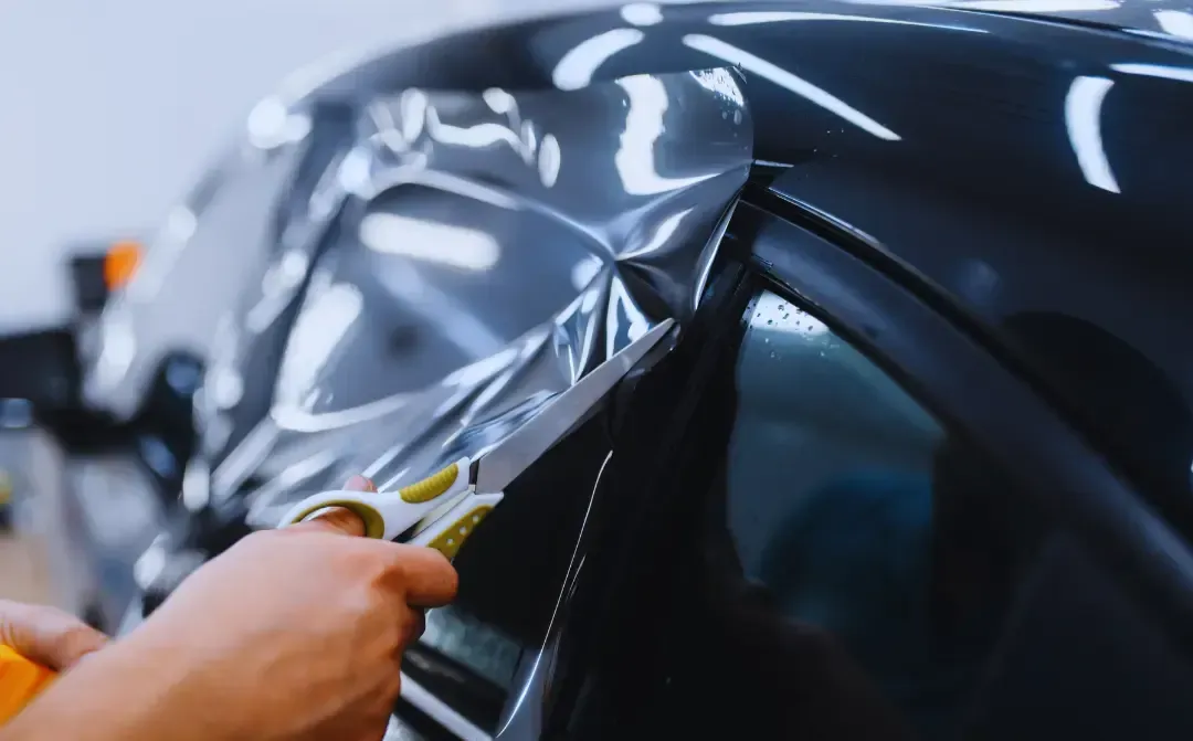 A person is cutting a piece of tinted glass on a car with scissors.