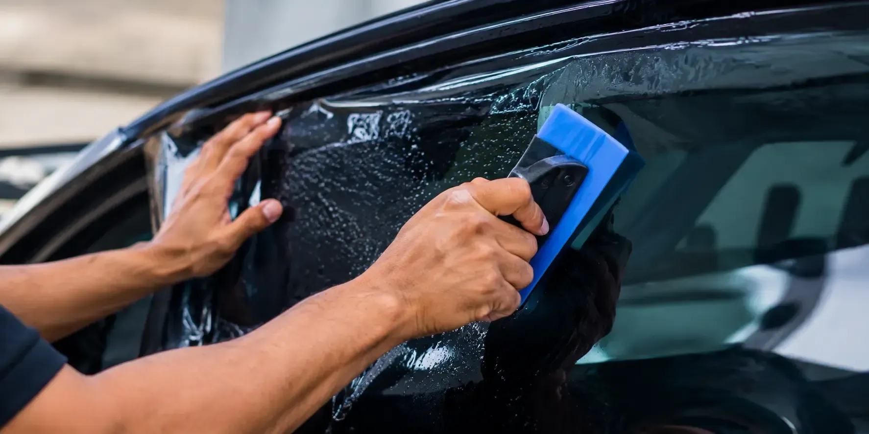 Close-up of a professional technician applying smooth, dark window tint to a car window using a squeegee tool.