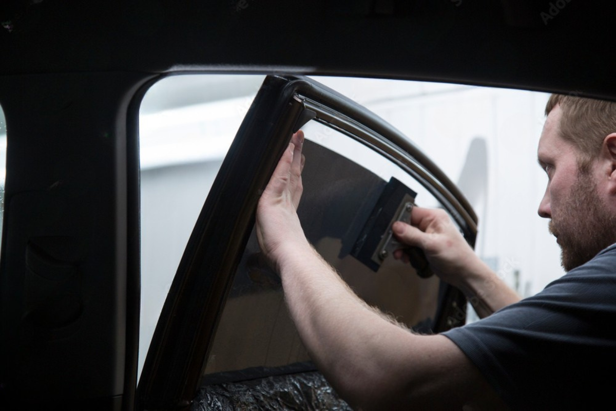 Man applying window tint to a car window with a squeegee, inside a garage.
