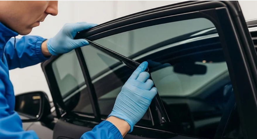 A person wearing blue gloves carefully applying a dark tint film to a car window.