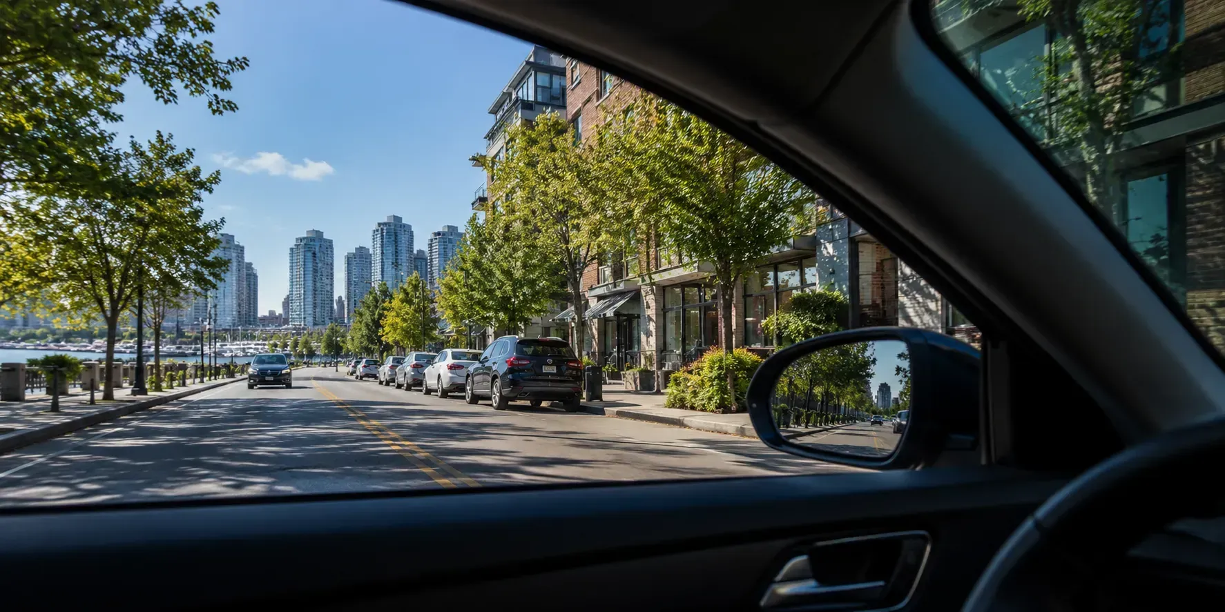 View from inside a car with tinted windows, showing clear visibility of a sunny urban street, parked vehicles, trees, and modern buildings outside.