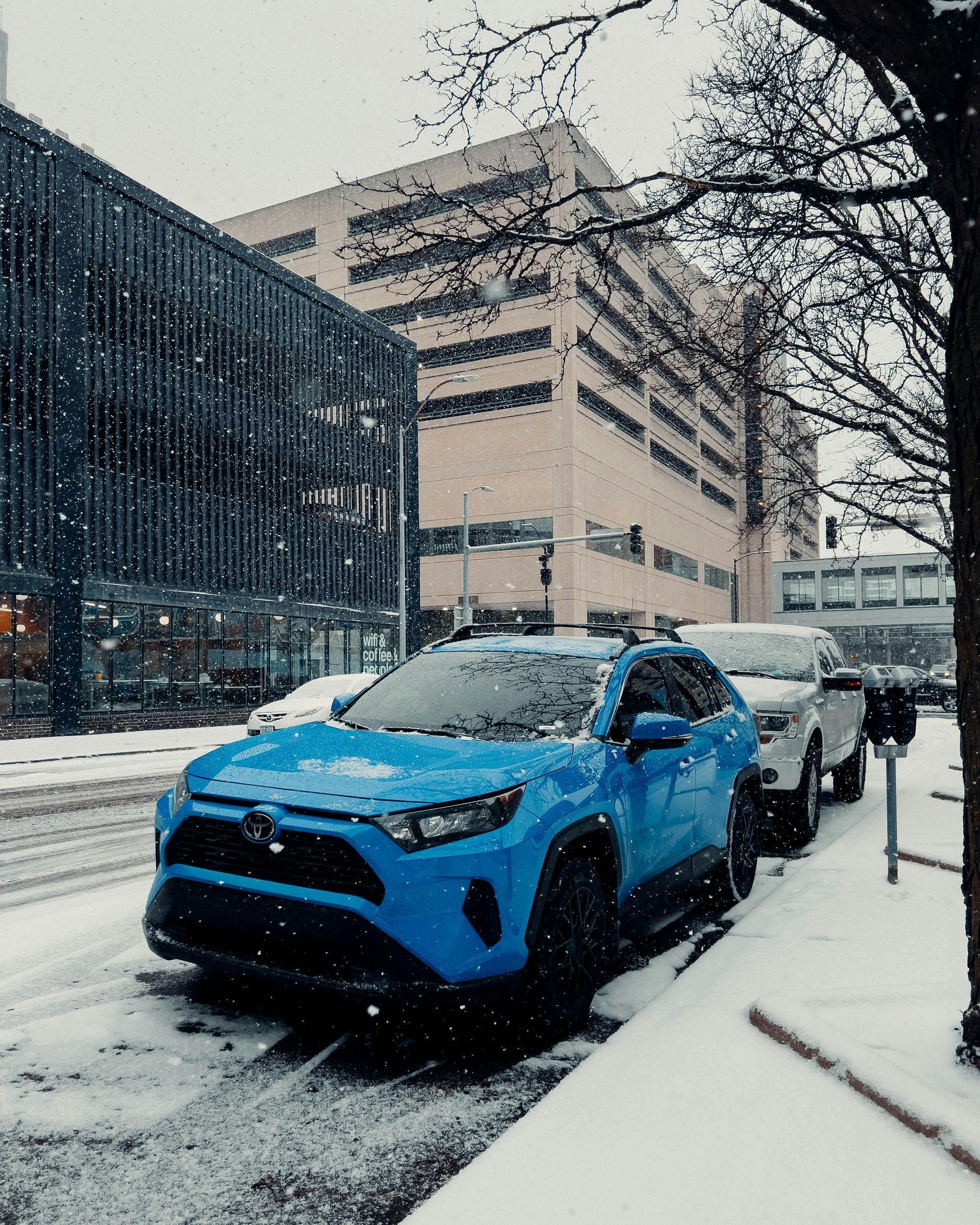 Blue car parked on a snowy city street beside tall buildings, with a person standing by the driver’s door.