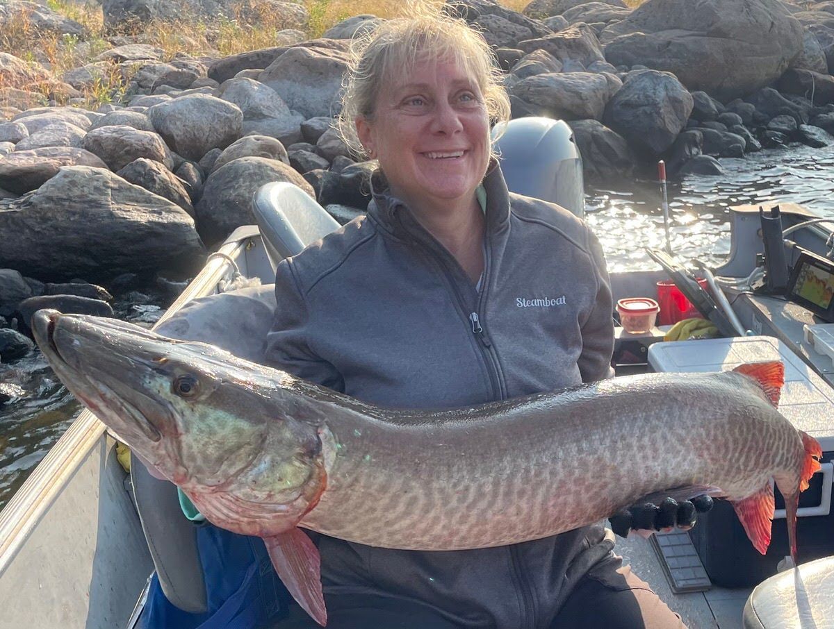 A woman is sitting in a boat holding a large muskie caught at Oak Lake.