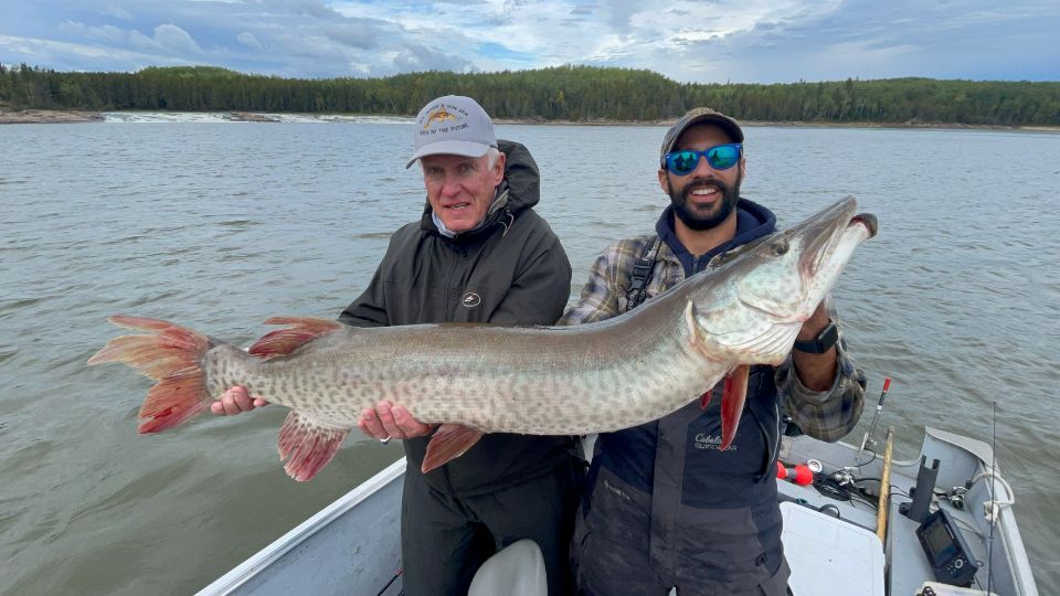 Two men are holding a large muskie in a boat on a lake.