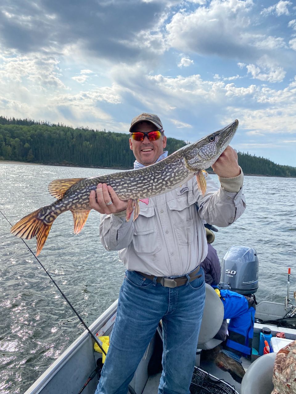 A man is standing in a boat holding a large Northern Pike.