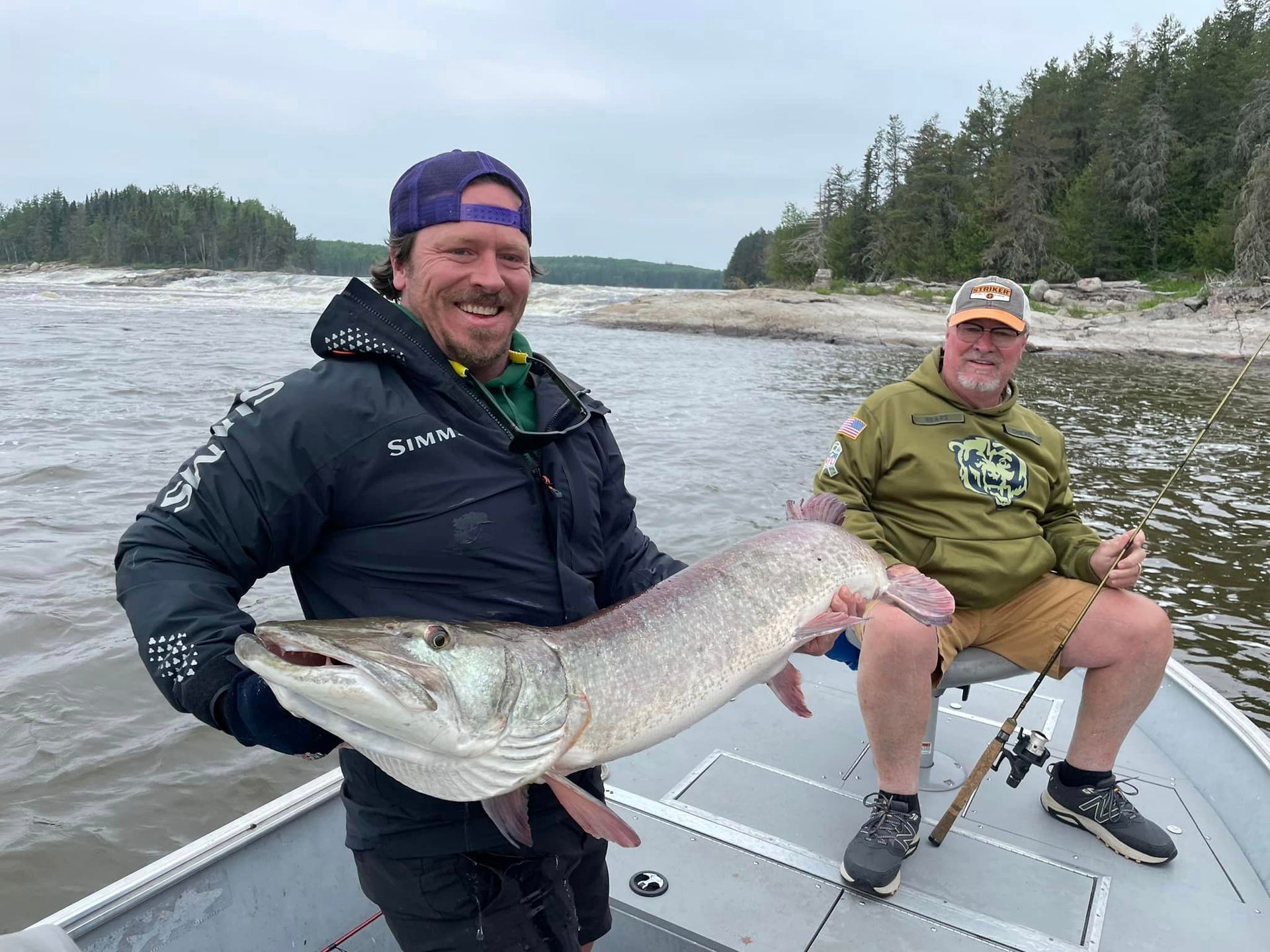 Two men are sitting on a boat holding a large muskie.