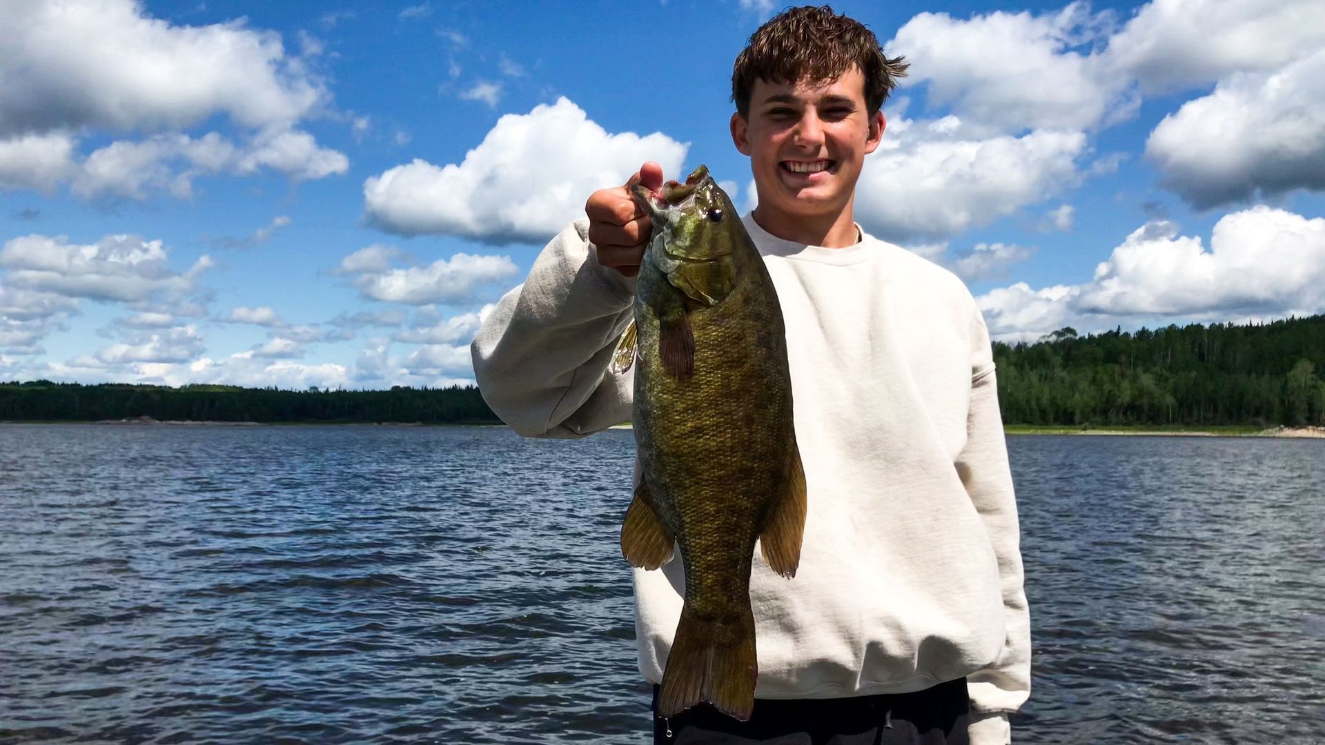 A young man proudly displays a smallmouth bass he caught, its glistening scales reflecting the sunlight, with a picturesque lake as the backdrop.