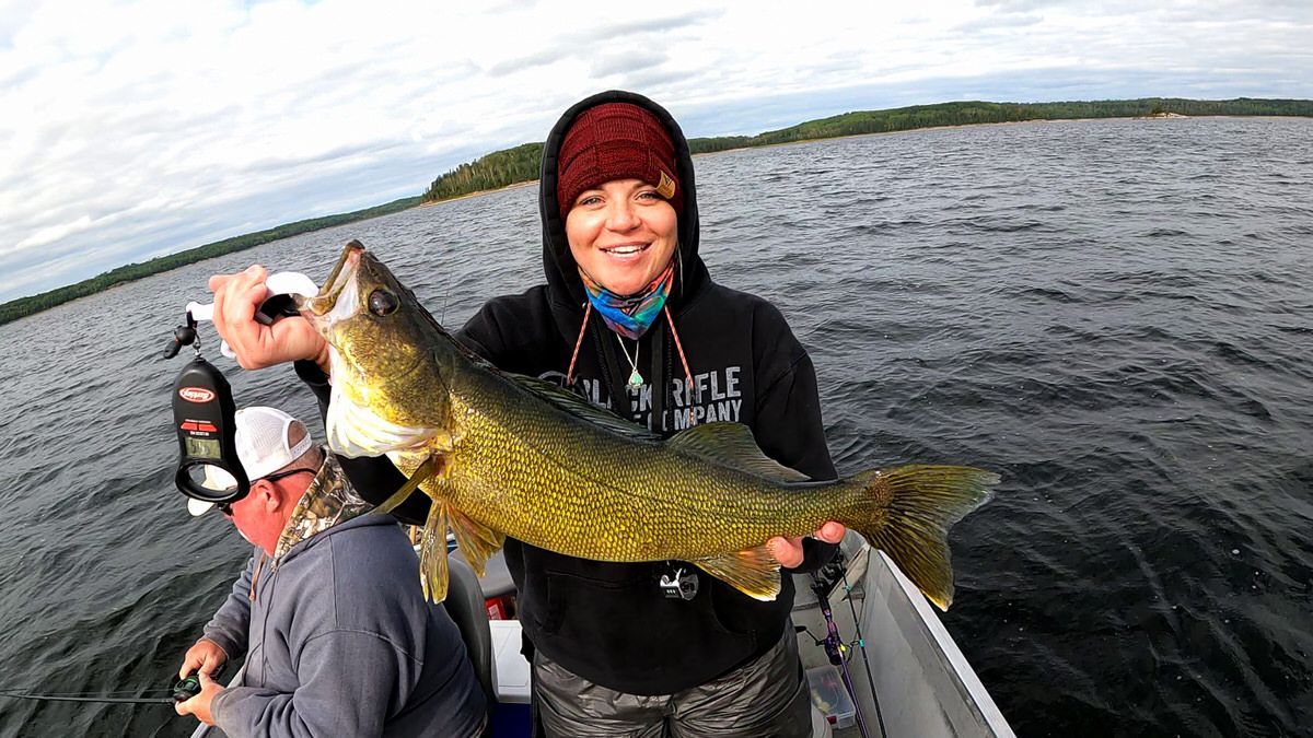 A woman is holding a large walleye fish in a boat on a lake.