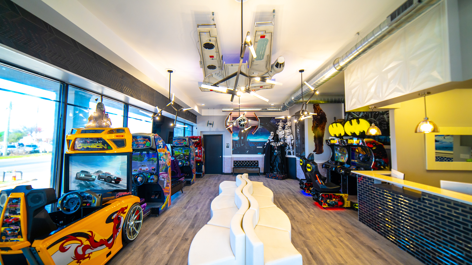 Colorful arcade with racing games and a central white track under bright ceiling lights.