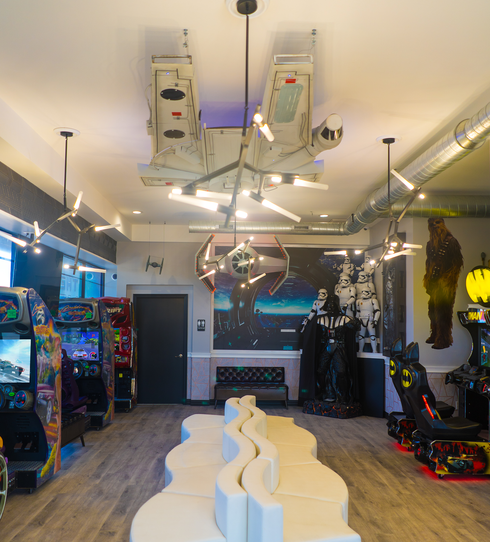 Colorful arcade room with pinball machines, ceiling-mounted ride, and a white wavy bench in the center