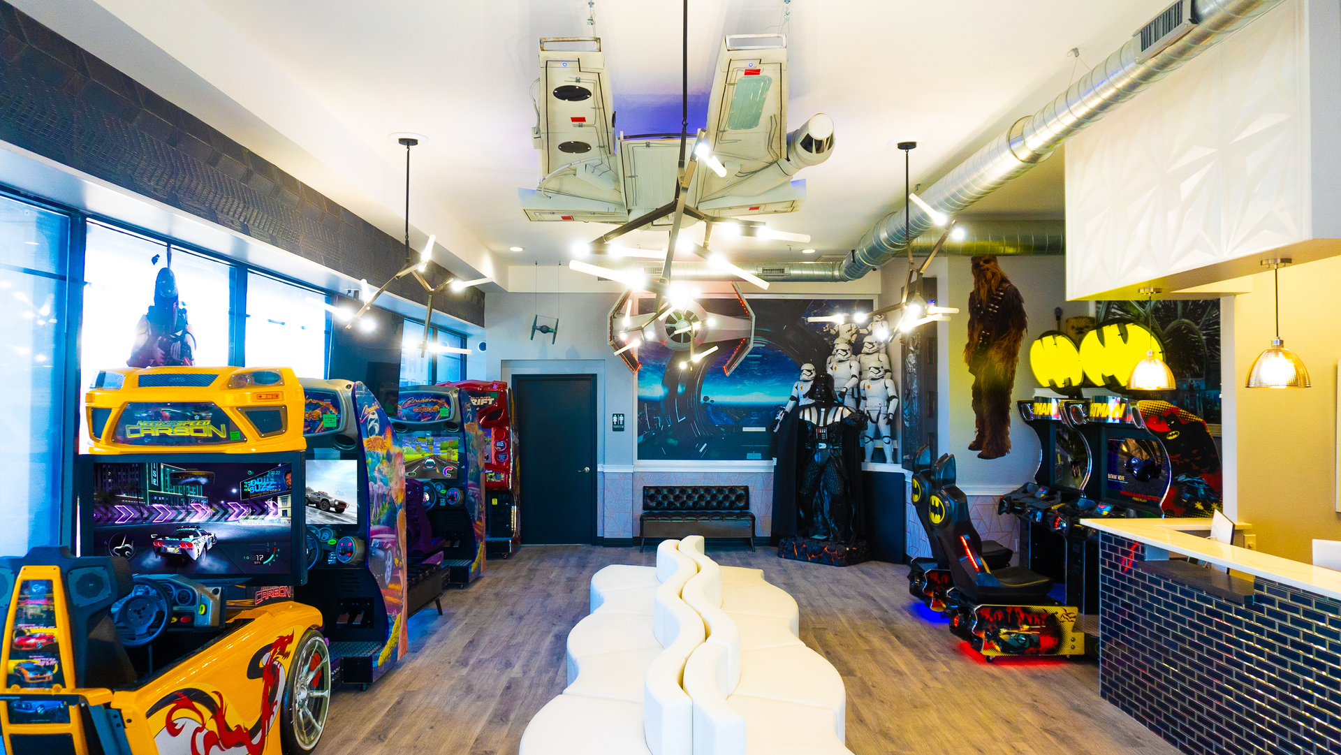 Bright game room with arcade cabinets, neon lights, hanging ceiling equipment, and a long white table center stage