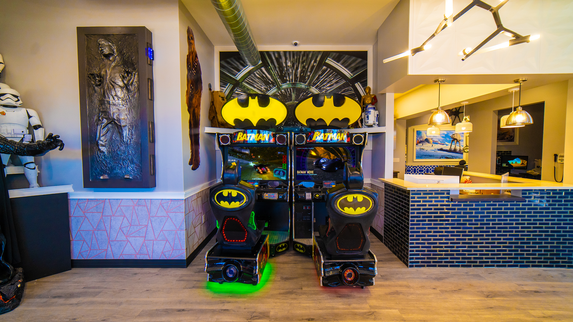 Batman-themed arcade racing machines in a brightly lit indoor game room, with a counter and seating area nearby