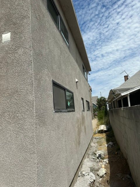 Narrow side yard between stucco house walls with a gravel path and cloudy sky overhead