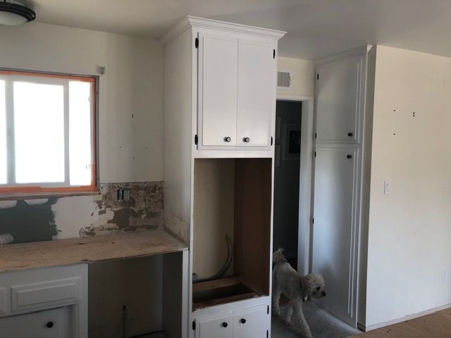 Small kitchen with white cabinets, countertop, window, open cabinet, and a dog standing near the doorway.