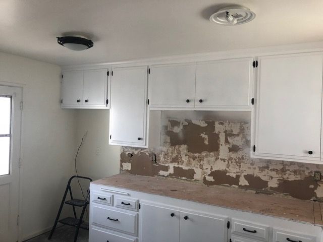 Small white kitchen with cabinets, stove area, and peeling wall paint during renovation.