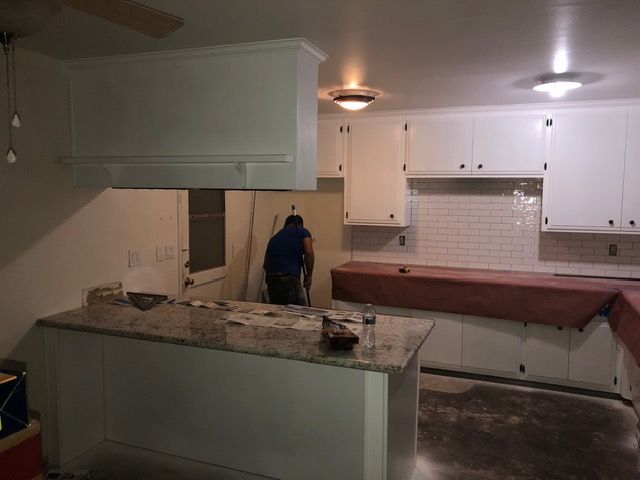 Kitchen under renovation with white cabinets, granite island, and a worker in blue near the back wall.