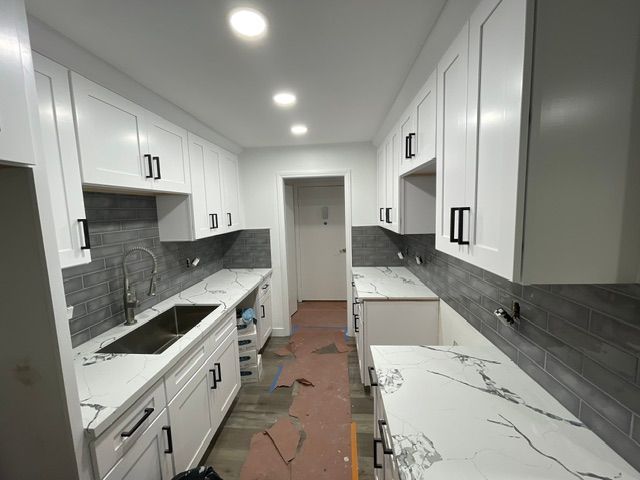 Bright white galley kitchen with marble counters, gray backsplash, and unfinished floor