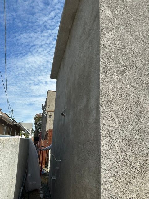 Narrow alley beside a stucco building under a blue sky, with utility wires and neighboring houses.
