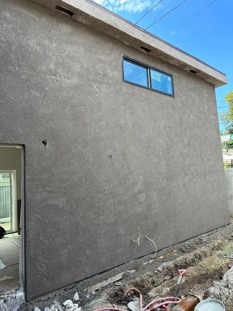 Gray stucco building exterior with a small blue window and unfinished ground beside it
