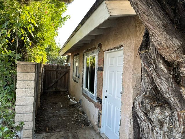 Narrow side yard beside a small house with a white door, window, and wooden gate under a tree.